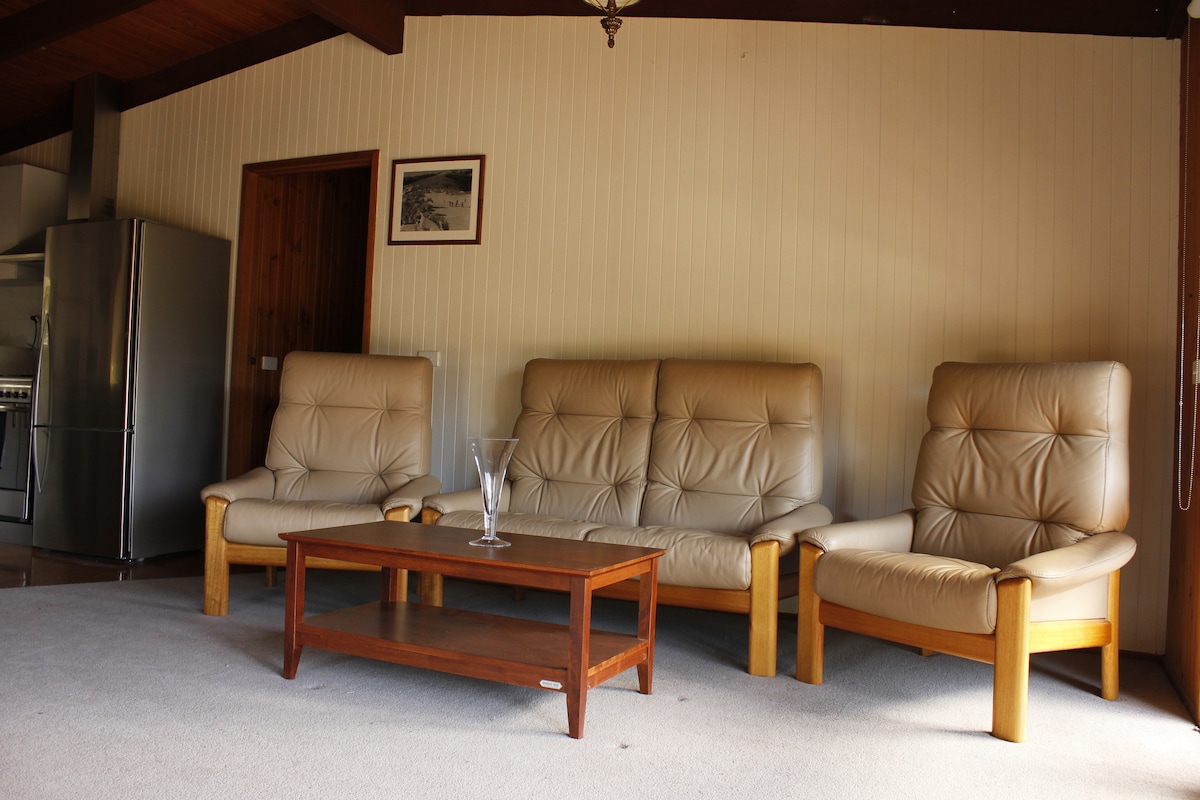 A comfortable living area is presented with two armchairs and a three-seater sofa, all featuring soft, neutral upholstery. A wooden coffee table is positioned in the center, accompanied by a decorative glass vase. The light-colored carpet and wooden beams add to the cozy ambiance.