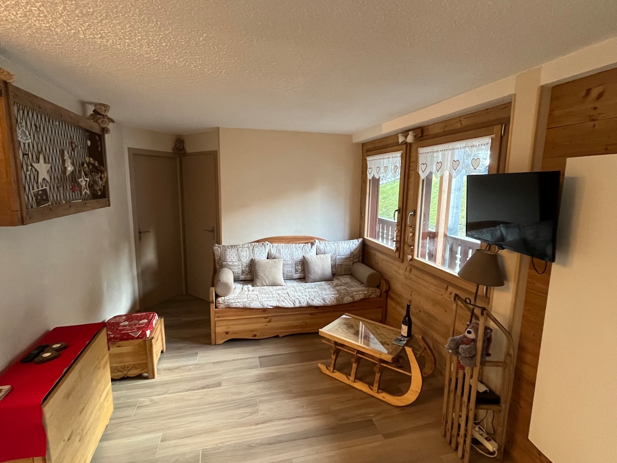 A modern living space features a wooden couch with decorative cushions, complemented by a glass coffee table. Natural light fills the room through large windows adorned with lace curtains. A television is mounted on the wall, and storage is provided by a wooden cabinet and a red table.