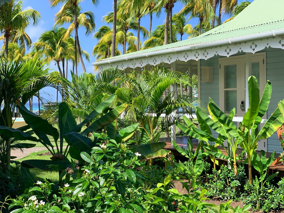 A charming villa is surrounded by lush tropical vegetation, featuring tall palm trees and vibrant green leaves. The structure's light blue exterior is complemented by decorative white trim, creating a serene connection with the natural landscape. Bright light filters through the foliage.