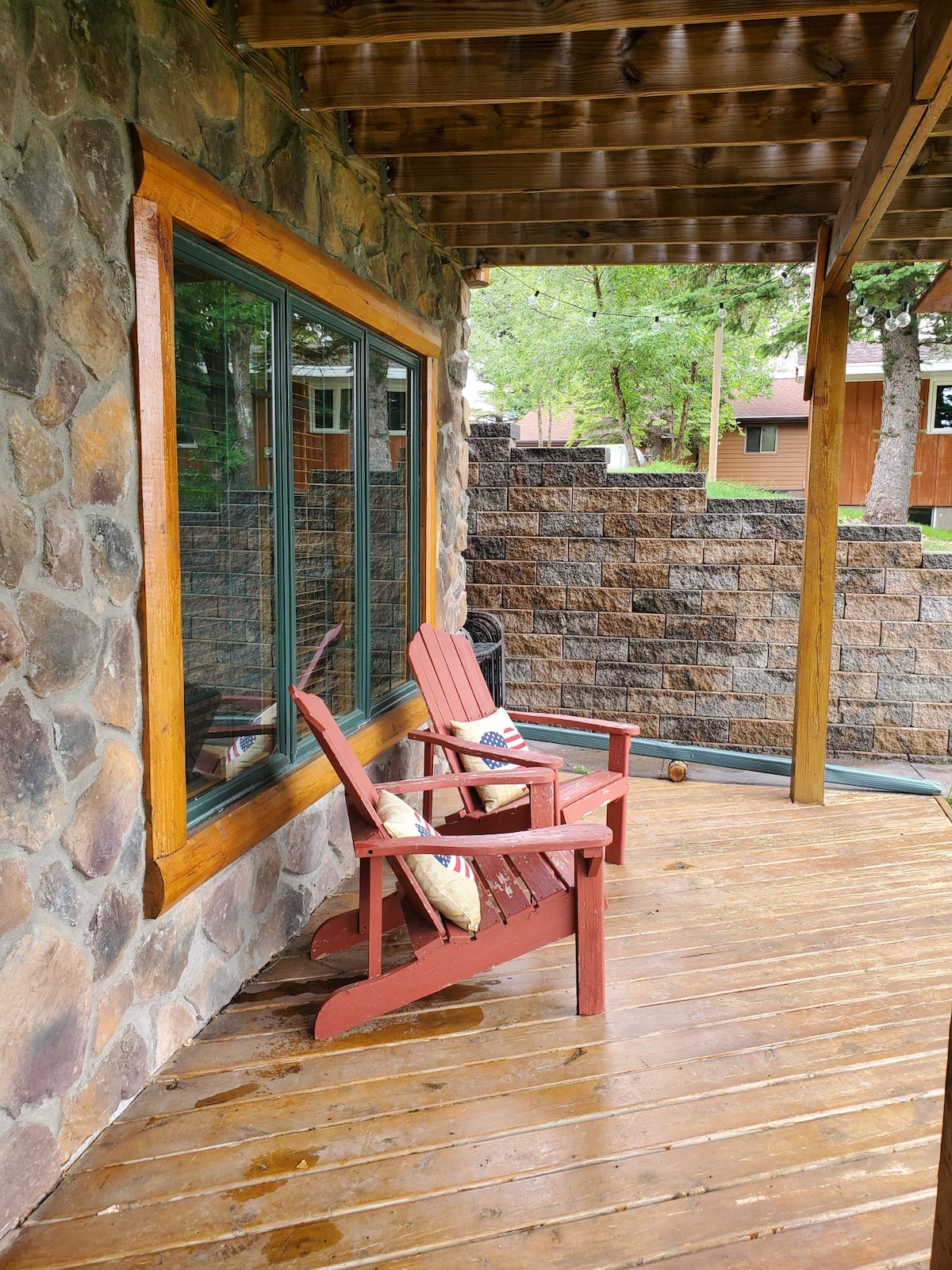 A shaded porch area features two red wooden chairs positioned on a wooden deck. Stone walls surround the space, with a large window allowing natural light to enter. A comfortable decorative pillow is placed on one chair, enhancing the relaxed setting.