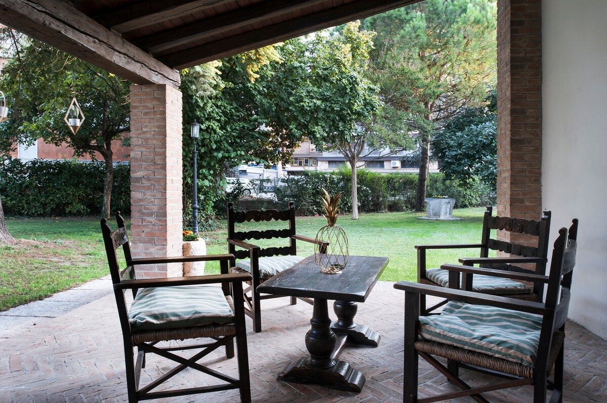 A covered porch area features a wooden table surrounded by four rustic chairs with outdoor cushions. Lush greenery surrounds the space, providing a tranquil setting for relaxation. Natural light filters through the trees, creating a welcoming atmosphere.