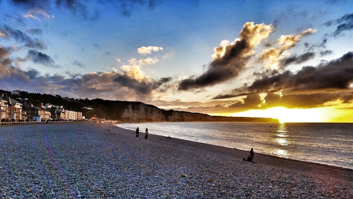 A beach scene captures the shoreline at sunset, with gentle waves lapping against the pebbled beach. Dark clouds contrast against the warm hues of the setting sun. Silhouettes of people can be seen walking along the water's edge, creating a serene atmosphere.
