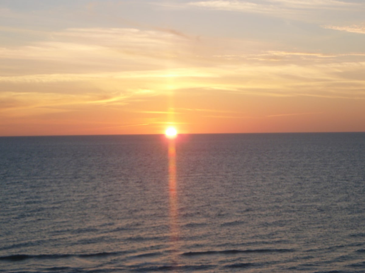 A serene sunset casts hues of orange and yellow over the water, reflecting softly on the gentle waves. The horizon displays a vivid sun descending into the Gulf, creating a peaceful and calming atmosphere.