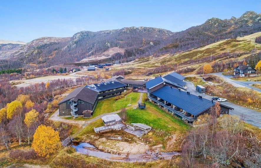 Aerial view of a large, modern building complex set in natural surroundings, featuring a mix of dark-toned wooden exteriors. Lush greenery and autumn foliage are visible, highlighting the hilly landscape in the background. Pathways weave through the grounds, leading to open spaces and a small stream.