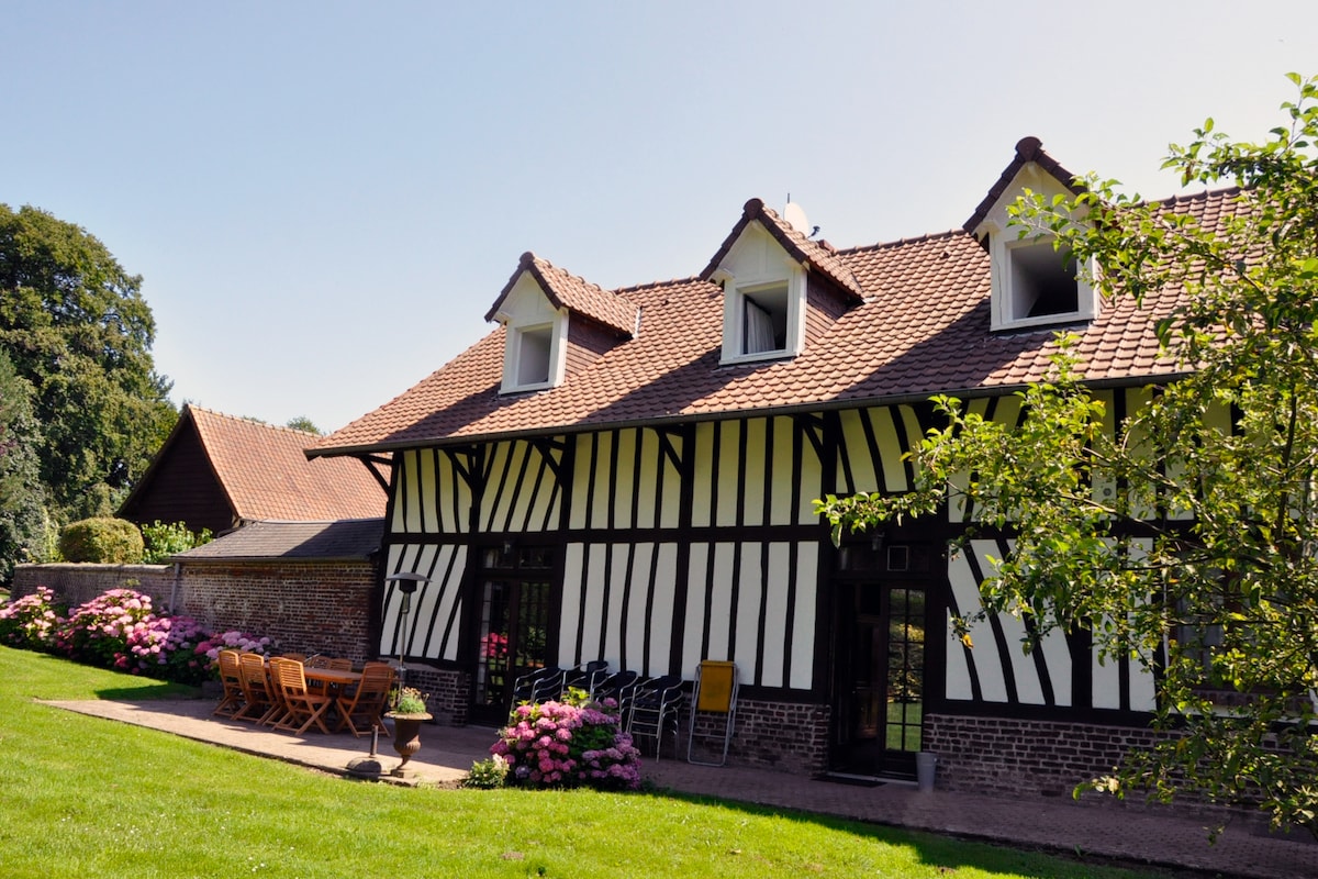 The exterior of a half-timbered house is presented, featuring a well-maintained garden with vibrant flower beds. A spacious terrace is furnished with a wooden dining set, surrounded by lush green grass and trees. The building showcases classic architectural details, enhancing its charming appearance.