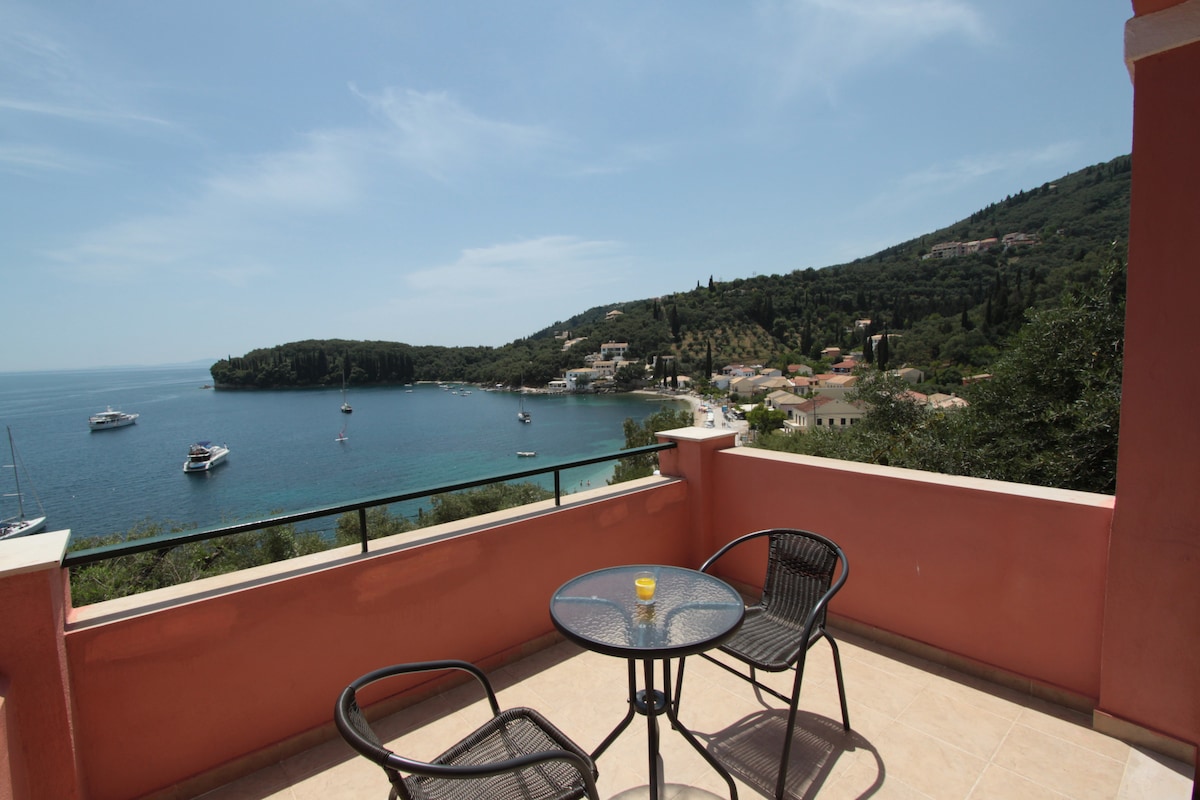 A balcony offers a clear view of the sea and surrounding hills. Two black chairs are positioned around a small glass table, while the calming blue water reflects the sky. Boats can be seen in the distance, enhancing the serene coastal atmosphere.