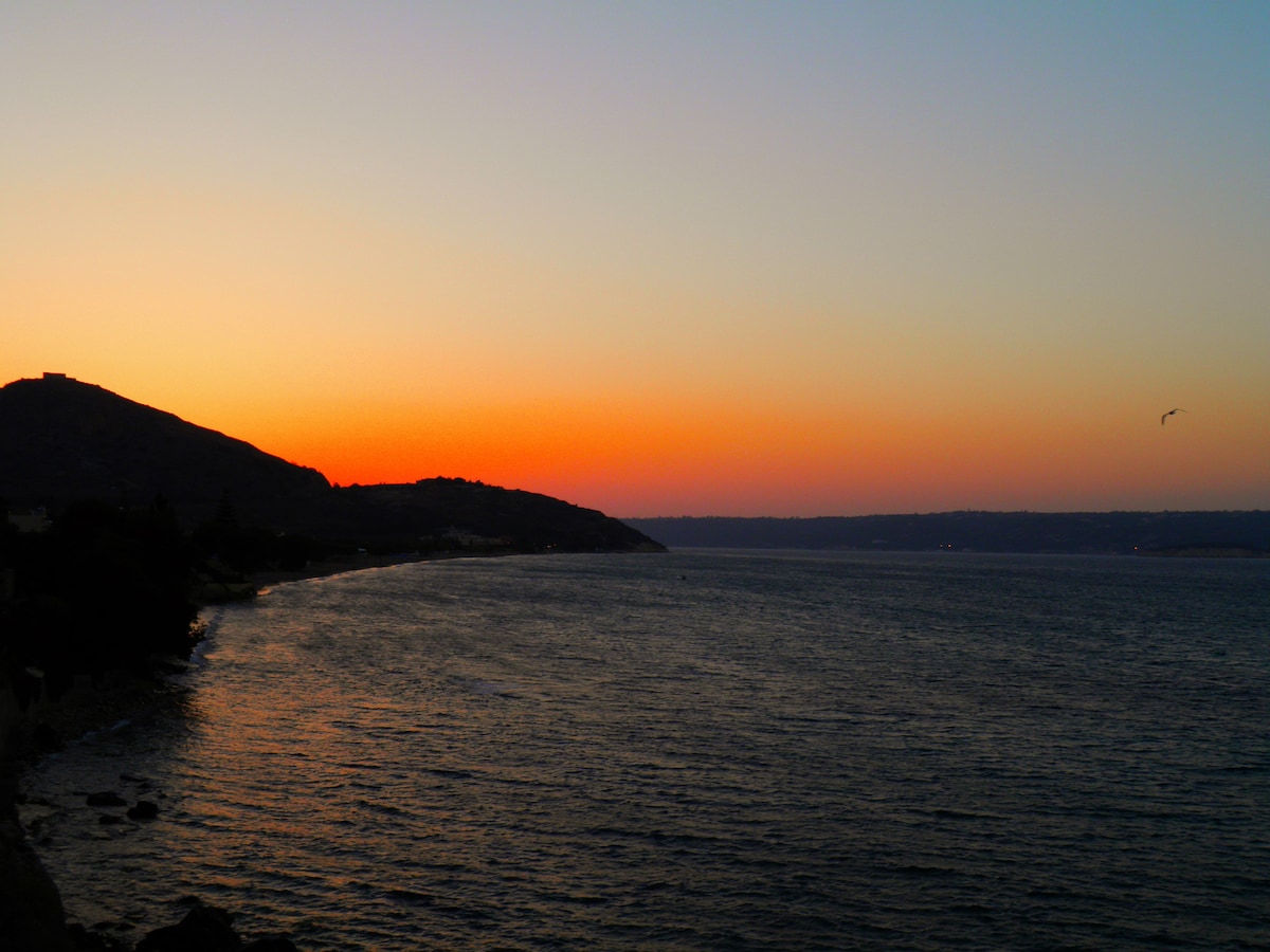 An expansive view of a coastline is captured during sunset, featuring warm orange and soft blue hues. The silhouette of a distant hill is visible on the left, while gentle waves create a serene atmosphere along the shoreline.