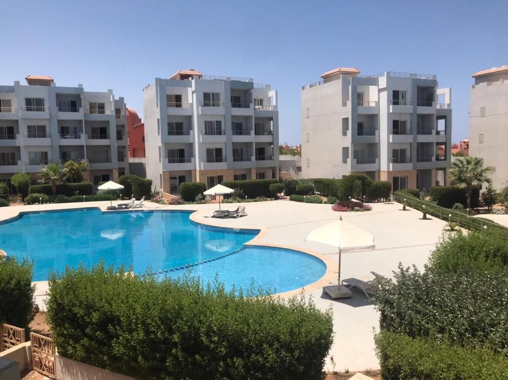A serene outdoor area features a large swimming pool surrounded by manicured gardens. Sun loungers and umbrellas provide shaded seating options. The backdrop includes modern multi-story buildings, and clear blue skies enhance the tranquil atmosphere.