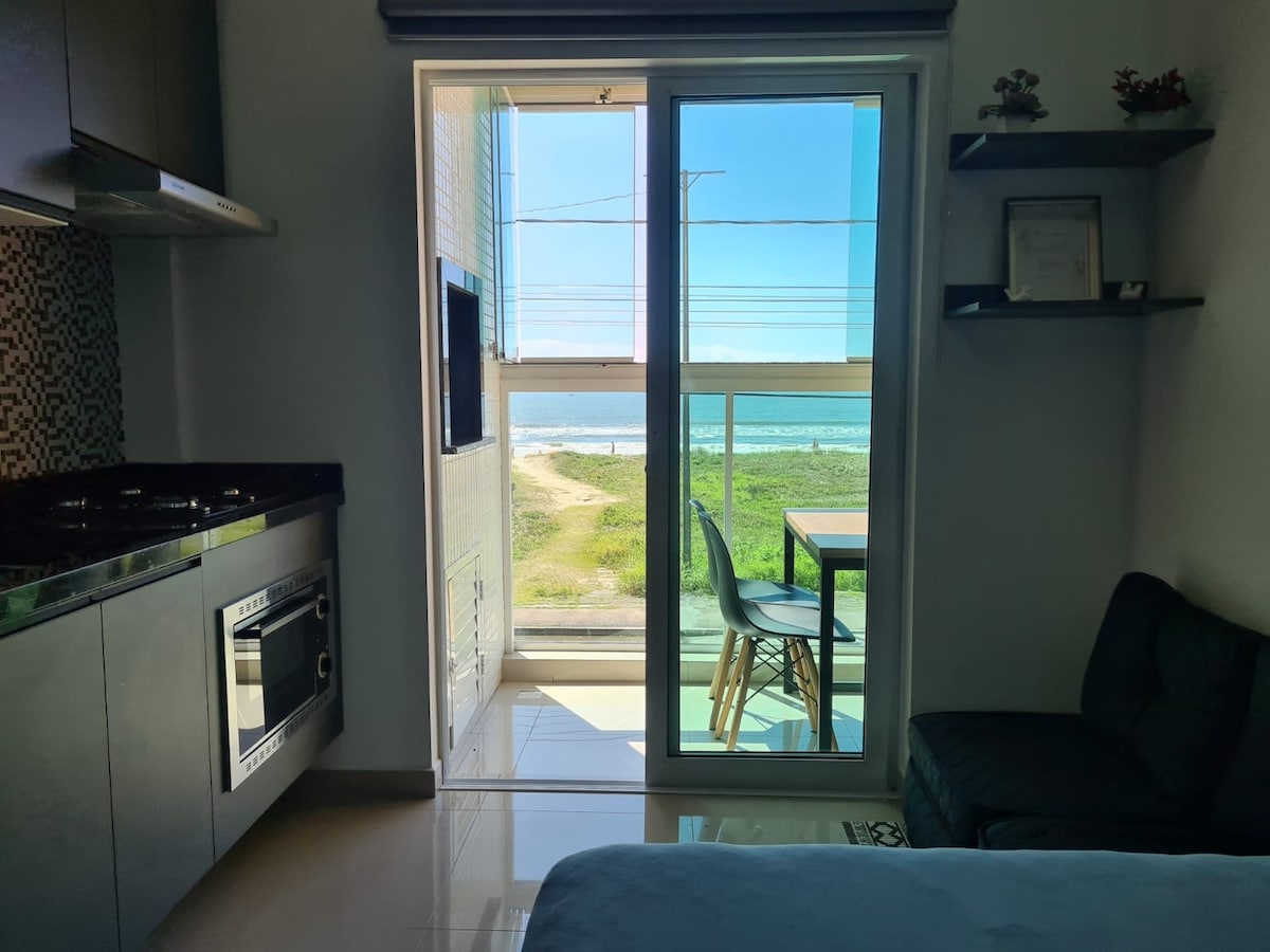 The image shows a light-filled studio with a view of the ocean through large sliding glass doors. A kitchenette is visible on one side, featuring dark cabinetry and appliances. A small dining table with chairs sits by the window, complementing the inviting space.