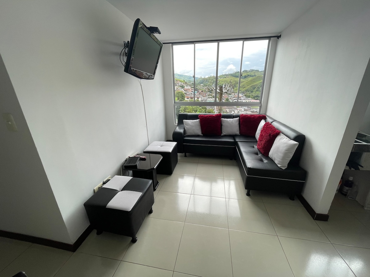 A compact living area features a black sectional sofa adorned with red and white cushions. A flat-screen television is mounted on the wall, and two small ottomans complete the space. Large windows offer views of the surrounding landscape.