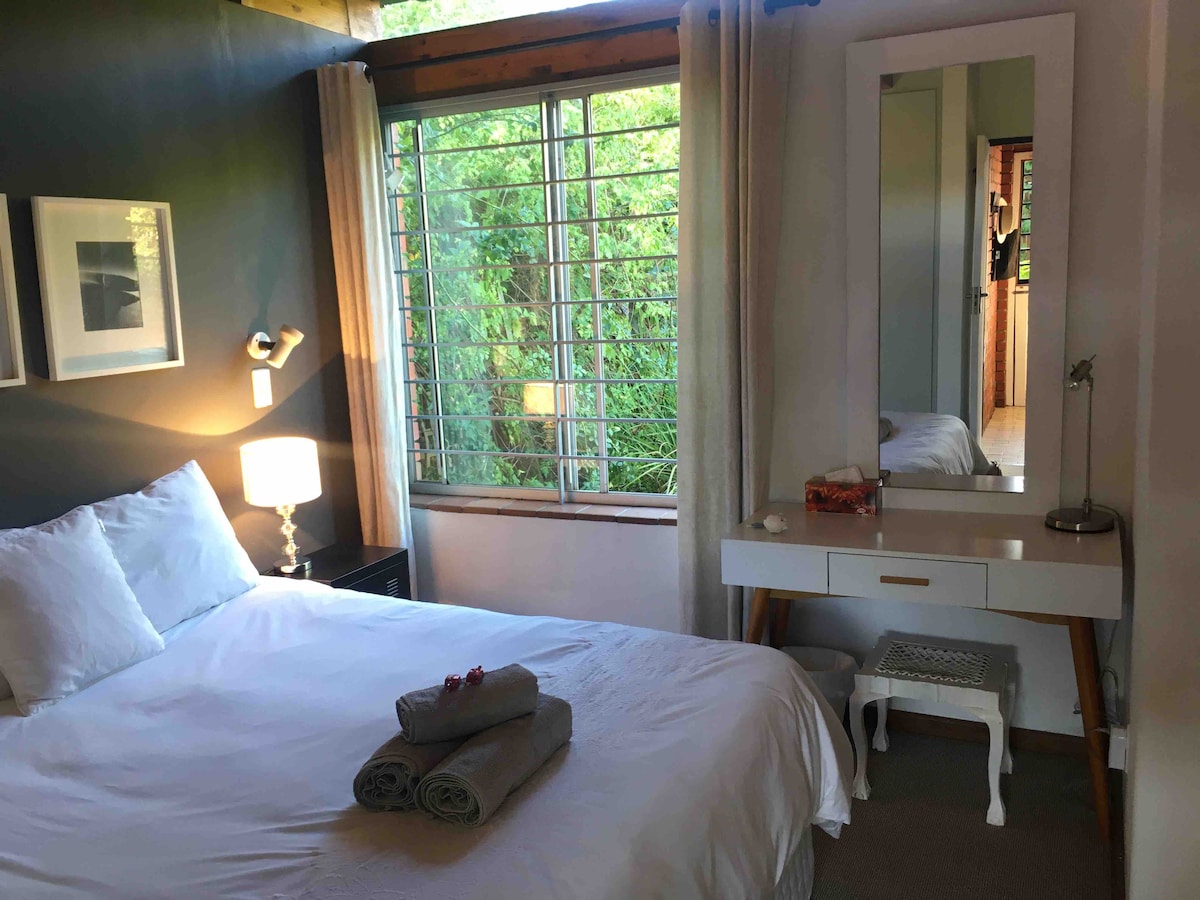 The bedroom features a spacious bed dressed in white linens, accompanied by neatly rolled towels. A bedside lamp provides soft lighting, while a large window with wooden slats allows natural light to fill the room. A small dressing table is positioned nearby.