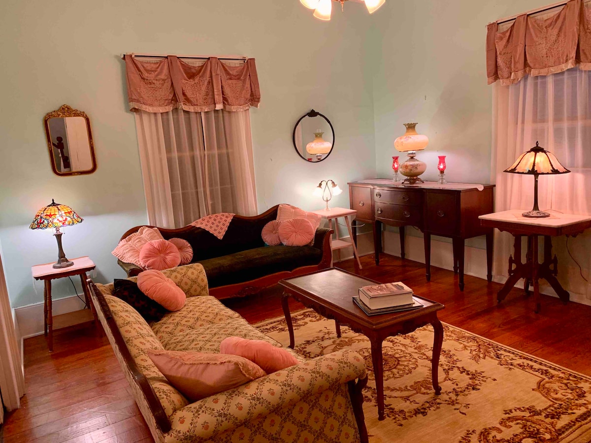 A tastefully decorated living room features vintage furnishings, including a floral settee and a dark sofa. Two delicate lamps illuminate the space, highlighting the hardwood flooring and a traditional area rug. Light green walls are adorned with decorative curtains, creating a warm ambiance.