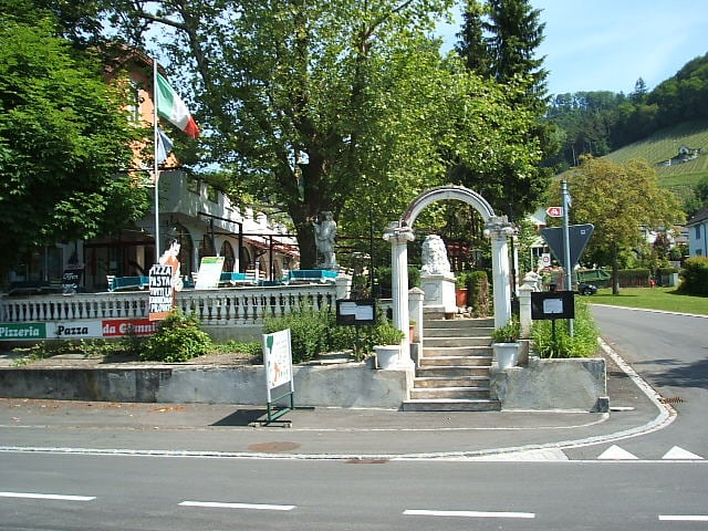 An entrance is framed by an archway and stairway, leading to a pizzeria with outdoor seating. Green trees provide shade, while decorative statues and potted plants add character. The setting is situated along a quiet road, complemented by a scenic vineyard backdrop.