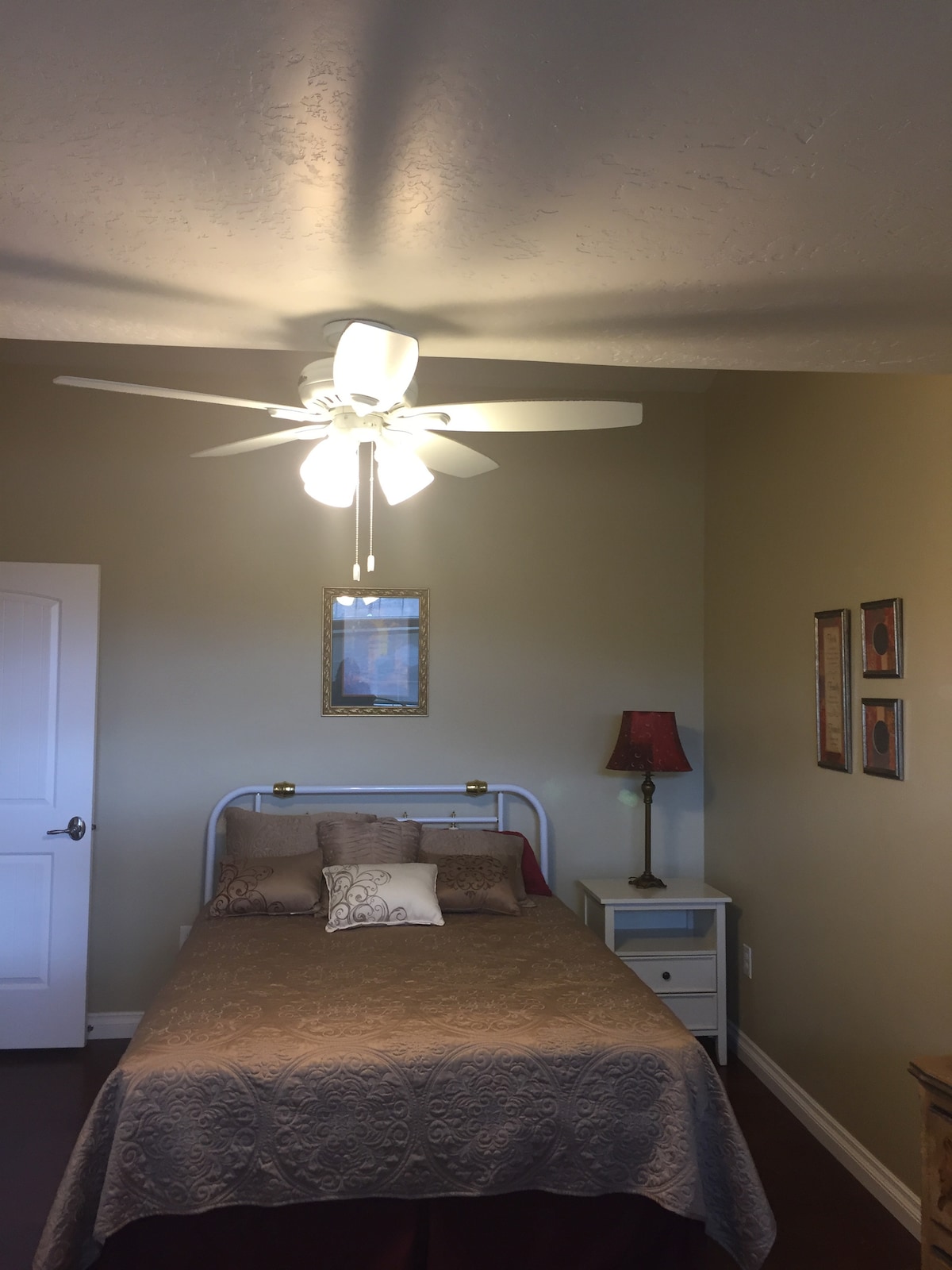 A cozy bedroom features a light-colored wall and a bed with a patterned coverlet and decorative pillows. A ceiling fan with five blades provides airflow, while a bedside lamp with a red shade offers soft lighting. Framed artwork adorns the walls.