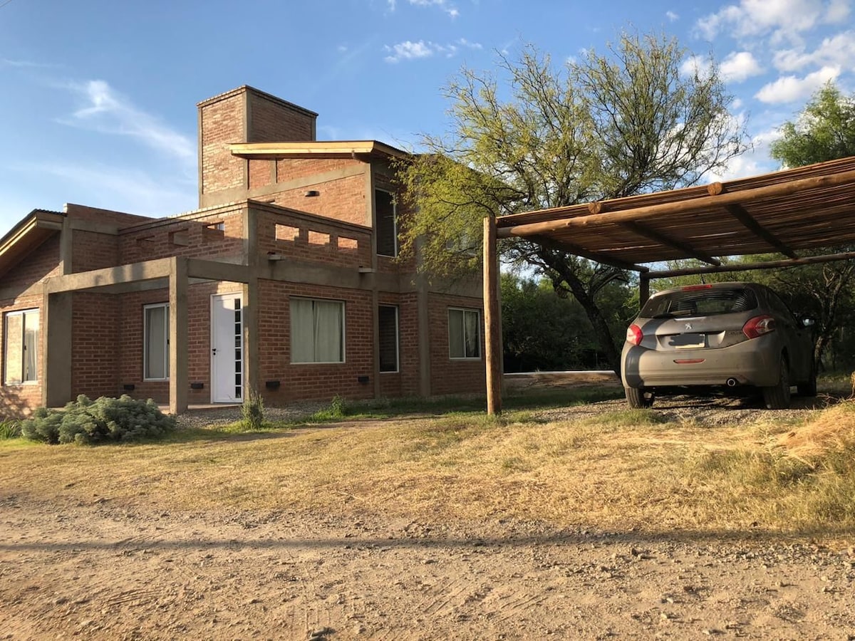 A two-story brick home is surrounded by natural greenery and open space. A covered parking area is visible beside the house, accommodating a vehicle. Large windows provide ample light, while a clear blue sky enhances the peaceful setting.