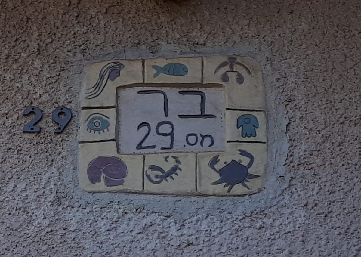 The exterior wall displays a decorative house number sign. The sign features a mosaic design with illustrated aquatic motifs surrounding the number 29. The colors are muted, combining earthy tones with subtle blues and greens.