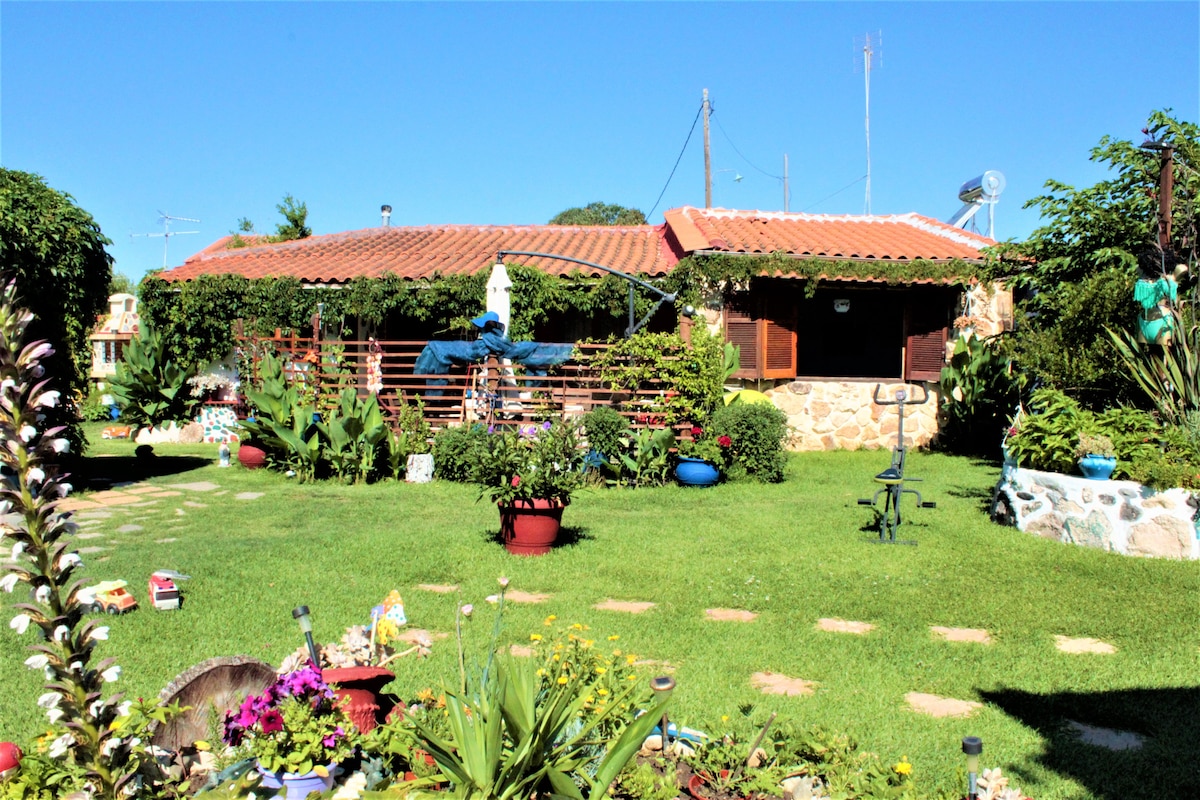 A vibrant garden surrounds the house, featuring a variety of colorful flowers and lush greenery. The stone house is complemented by a terracotta roof and wooden window shutters. Pathways of stones lead through the garden, creating a welcoming outdoor space.