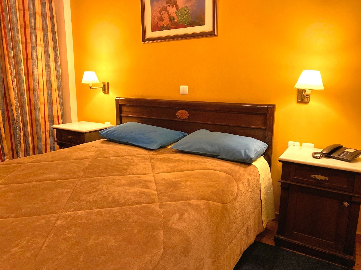 A spacious bed with a brown patterned blanket and blue pillows is situated against a warm yellow wall. Soft lighting from wall sconces creates a gentle glow. Two bedside tables are positioned beside the bed, and colorful striped curtains frame the window.
