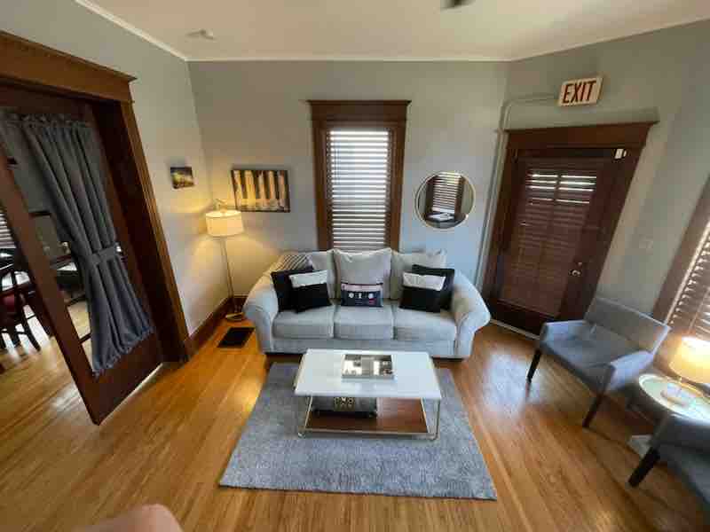 The living room features a light gray sofa with decorative pillows, arranged around a sleek coffee table atop a soft area rug. Natural light filters through the window blinds. A lamp illuminates the corner, and an exit sign is visible on the wall.