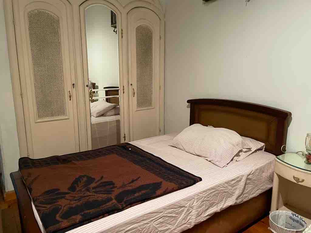 A spacious bed is positioned against a light-colored wall, accompanied by a neatly arranged blanket. A mirrored wardrobe reflects the room's soft lighting, adding depth to the space. A bedside table holds small decorative items, contributing to the room’s serene atmosphere.