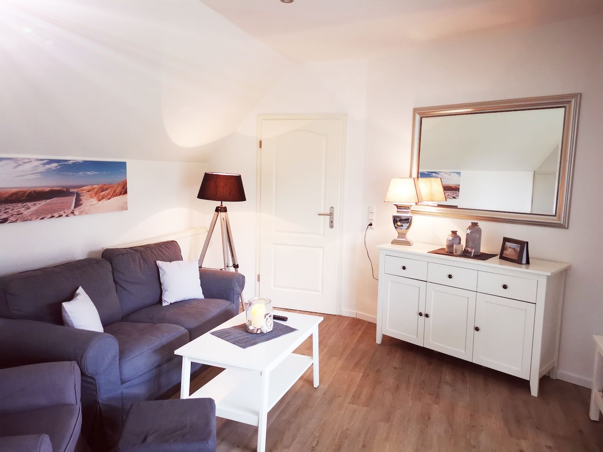 A modern living area features a comfortable gray couch adorned with white pillows and is accompanied by a small white coffee table. A simple side table with a lamp and a decorative mirror add to the space. Natural light illuminates the room, enhancing its inviting ambiance.