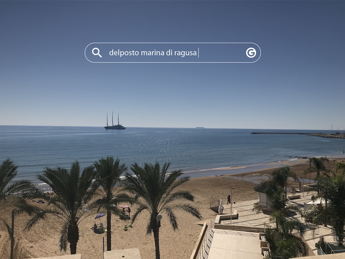 A serene beach scene is displayed, highlighting soft sandy shores meeting calm blue waters. Palm trees frame the foreground, while a sailing ship is visible on the horizon under a clear blue sky. The tranquility of the setting is inviting for beachgoers.