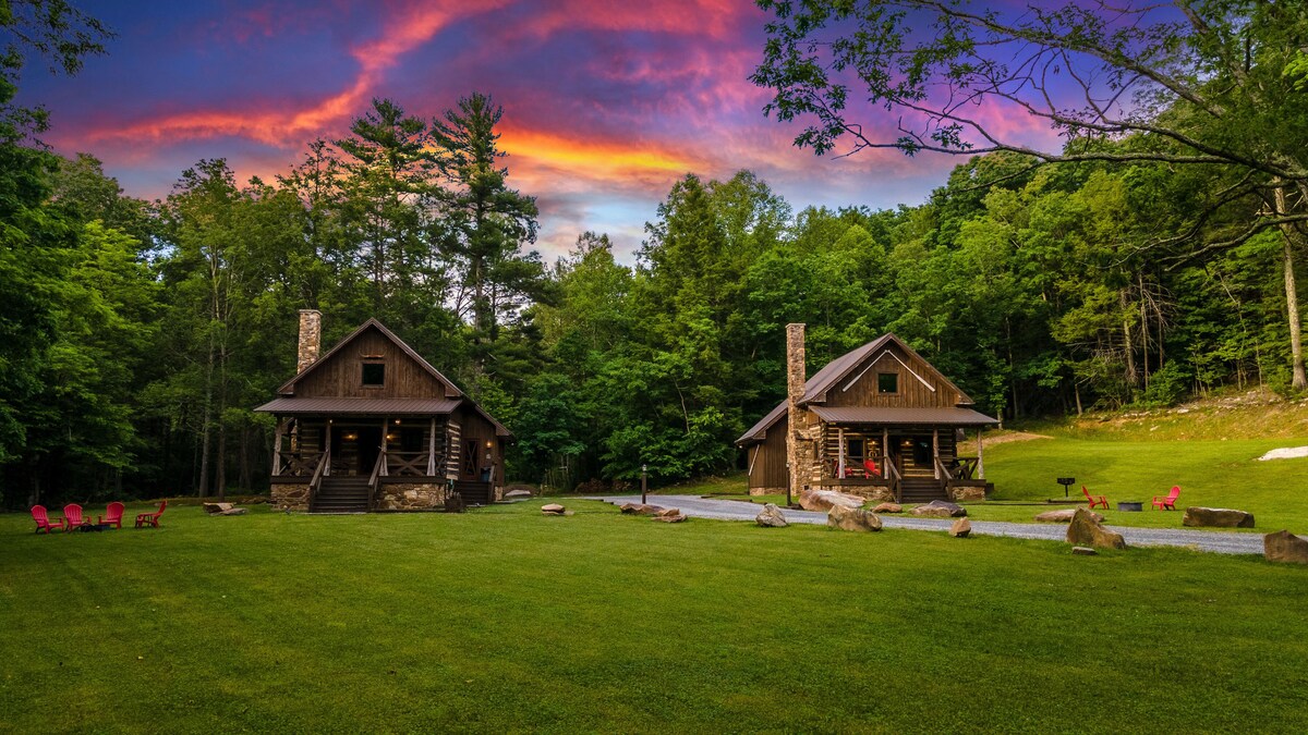 The image captures two rustic cabins set within a lush green landscape, surrounded by tall trees. A vibrant sunset sky paints the backdrop, displaying shades of purple and orange. A grassy area lies in the foreground, featuring natural seating and a gravel pathway leading to the cabins.