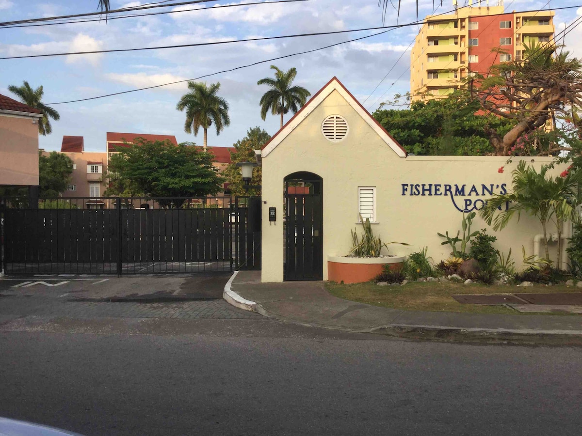 A gated entrance is depicted, featuring a clean, white wall with the text 'Fisherman's Point' prominently displayed. Lush greenery fills the surrounding area, with palm trees visible in the background. The entrance is flanked by a neatly maintained garden, and a black gate can be seen partially open.