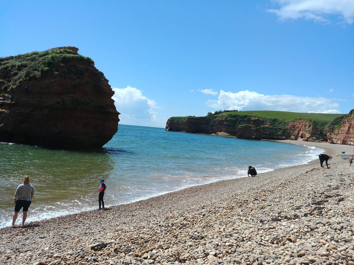 A scenic beach scene features pebbles along the shore, with gentle waves lapping at the water's edge. Individuals are seen exploring the shoreline while a large rock formation rises prominently from the sea. Lush green cliffs frame the background under a clear blue sky.