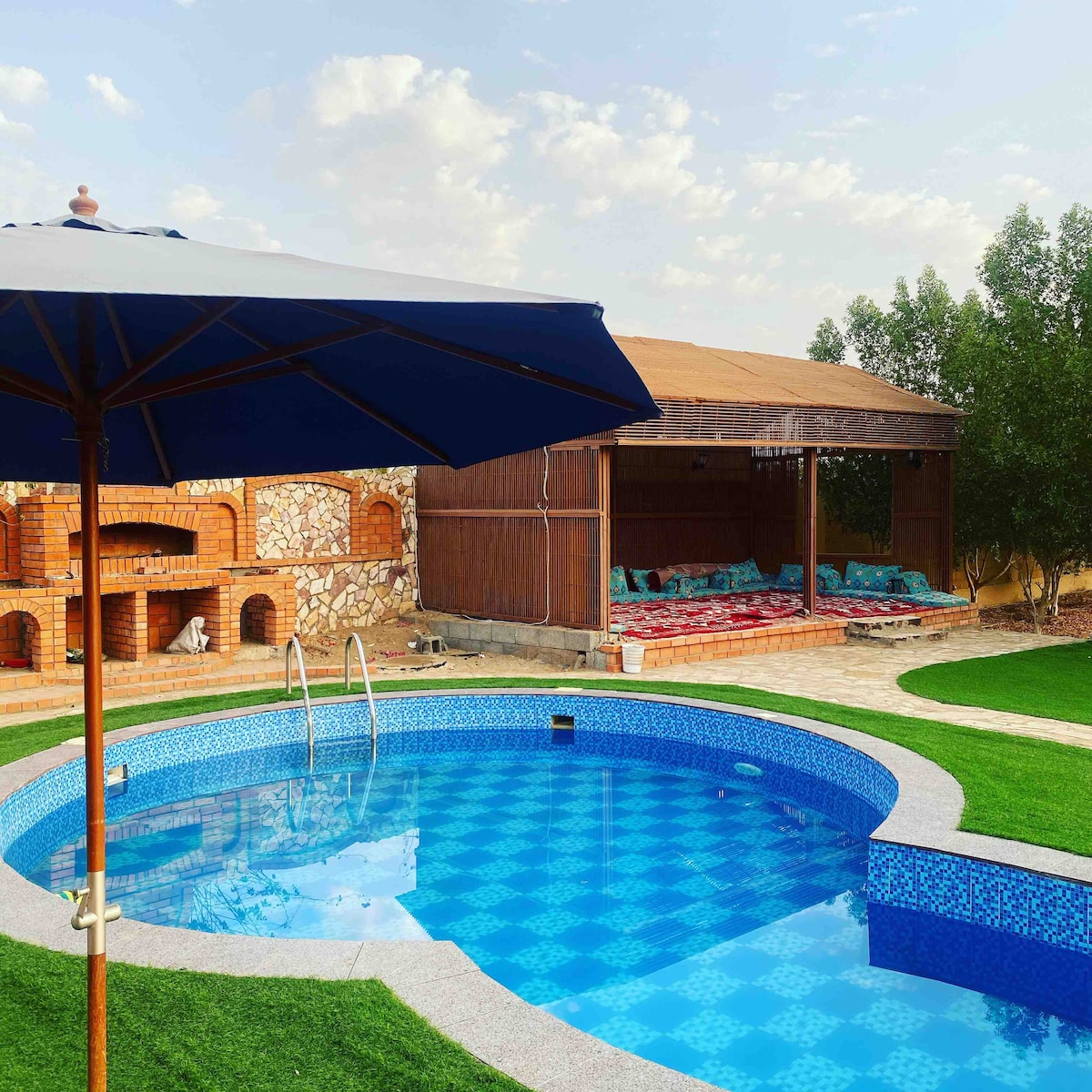 A refreshing pool is surrounded by manicured green grass and a shaded area with traditional seating. An umbrella provides shade near the pool's edge, while the stone structure in the background features a barbecue area. Scenic skies add to the serene environment.