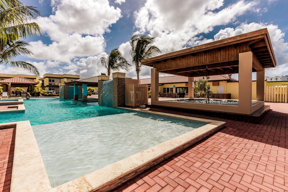 A large swimming pool is set in a spacious outdoor area, complemented by palm trees. A shaded gazebo provides a relaxing spot, while the pool features smooth tiles and gentle water ripples. The surrounding area is paved with red bricks, enhancing the inviting ambiance.