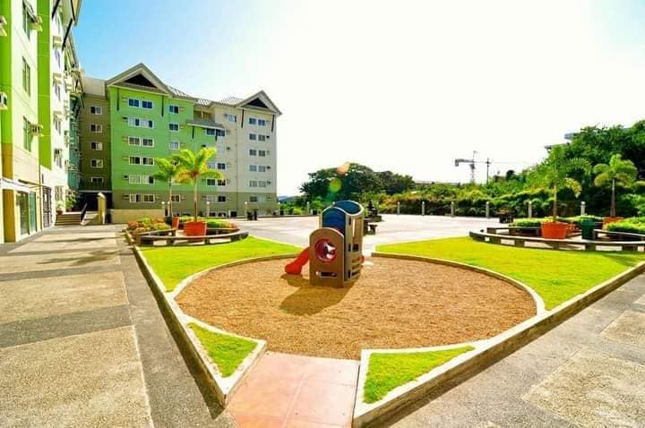 The outdoor area features a well-maintained, grassy playground surrounded by colorful flower beds. A children's play structure with a slide and climbing features is positioned in the center, providing a cheerful spot for recreation. The surrounding buildings reflect a modern architectural style under a bright blue sky.