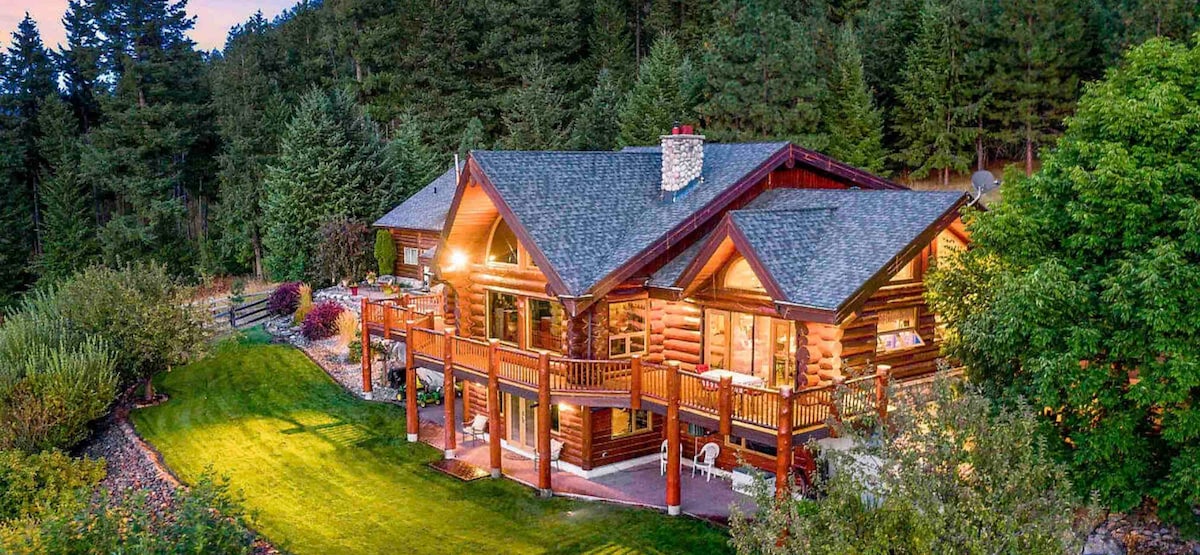 The log home is surrounded by lush greenery and showcases warm lighting that highlights its wooden structure. Multiple decks are visible, perfect for enjoying outdoor moments. The expansive lawn leads to a scenic backdrop of trees, enhancing the tranquility of the setting.