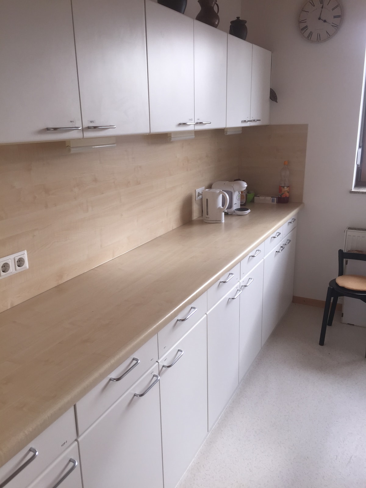 A bright kitchen area features a smooth, light-colored countertop with white cabinetry. Essential appliances, including a kettle and coffee machine, are visible, alongside a small selection of utensils. Natural light enters the space from a window, complemented by a clock mounted on the wall.