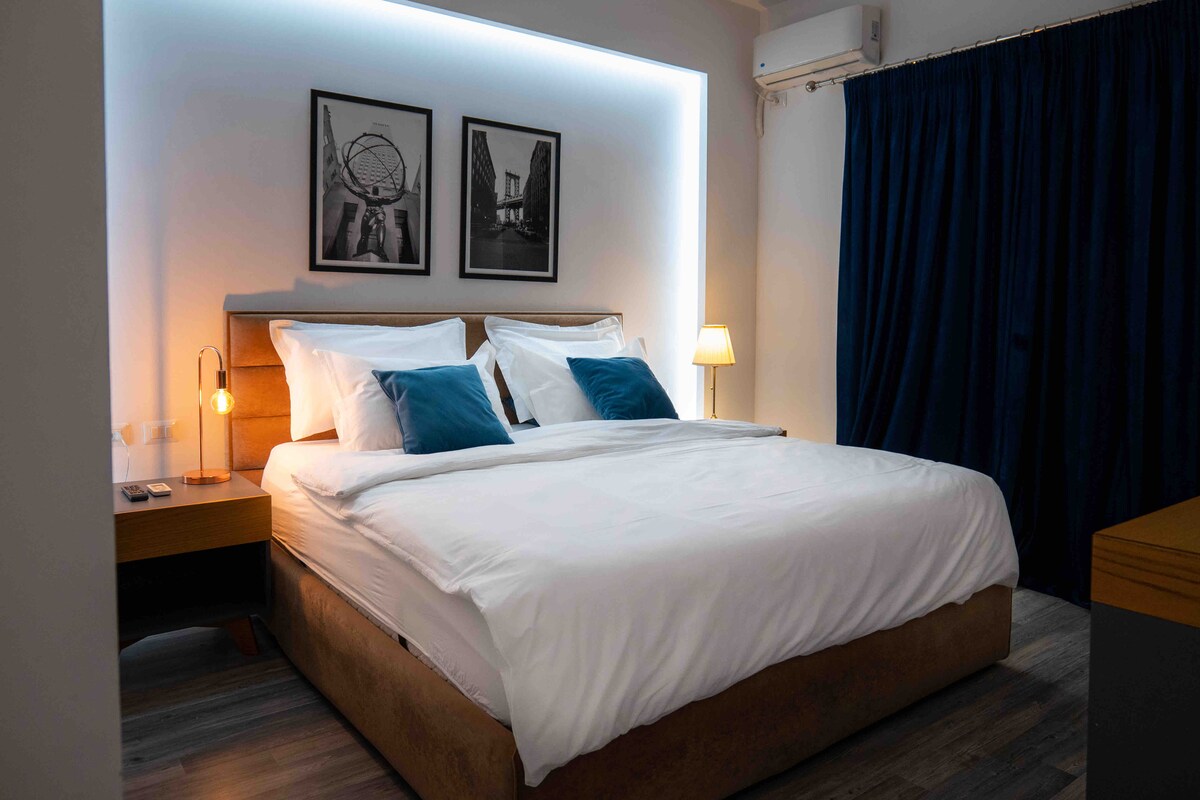 A spacious bed with white linens and plush pillows is centered in the room. Two framed black-and-white photographs decorate the wall above the bed. A soft bedside lamp emits warm light, complementing the dark curtains that frame the window.
