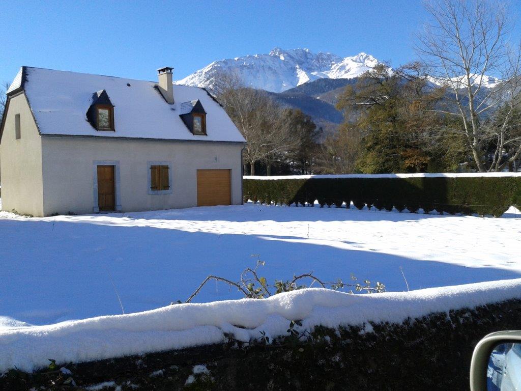 A snow-covered house with a sloped roof is positioned in front of a mountainous backdrop. The surrounding lawn, blanketed in white snow, contrasts with the greenery of trimmed hedges and trees. Clear skies reflect a serene winter atmosphere.
