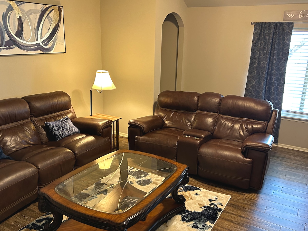 A comfortable living area features two brown leather sofas, with one containing a built-in cup holder. A wooden coffee table with a glass top rests on a patterned rug. A lamp provides soft lighting, and a window is adorned with decorative curtains.