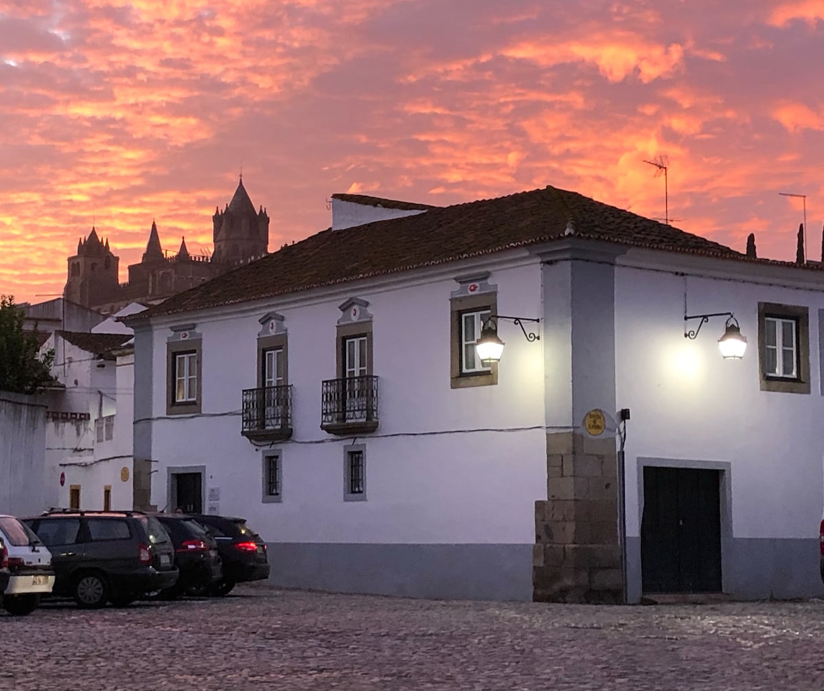 A historic white building is illuminated by soft electric lights against a vibrant sunset sky, creating a warm evening ambiance. Two balconies are visible on the upper floor, and the cobblestone street is lined with parked cars, adding to the charm of the scene.