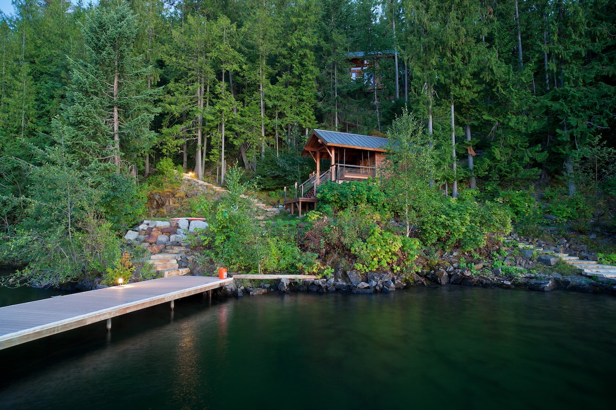 A wooden dock extends into the calm waters, leading to a charming cabin nestled among lush greenery. The structure features a sloped roof and a welcoming porch, surrounded by trees that enhance the serene lakeside environment.