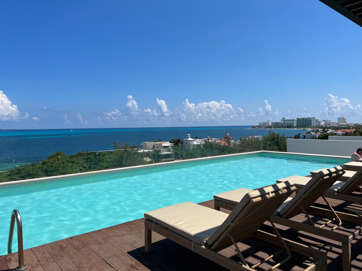 An inviting infinity pool extends over a wooden deck, offering stunning views of the vibrant blue sea and distant shoreline. Sun loungers line the edge, providing a relaxing spot to enjoy the scenic horizon and clear skies.
