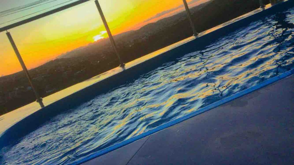 A tranquil pool is visible at sunset, with water reflecting vibrant hues of orange and yellow. The pool is located on a balcony, allowing for a panoramic view of the surrounding landscape and city below.