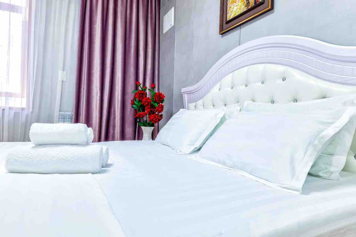 A spacious bed with crisp white linens is positioned against a light grey wall, complemented by patterned throw pillows. A bouquet of red flowers in a white vase adds a touch of color. Sheer curtains allow natural light to filter softly into the room.