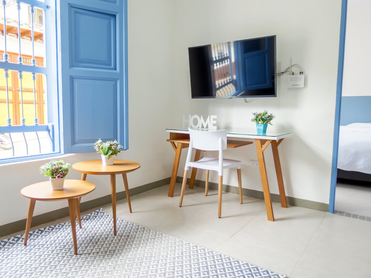 A light and airy living space features a modern desk with a chair and a mounted television. Two small wooden tables sit on a patterned rug, accompanied by potted plants. A window with blue shutters allows natural light to fill the room.