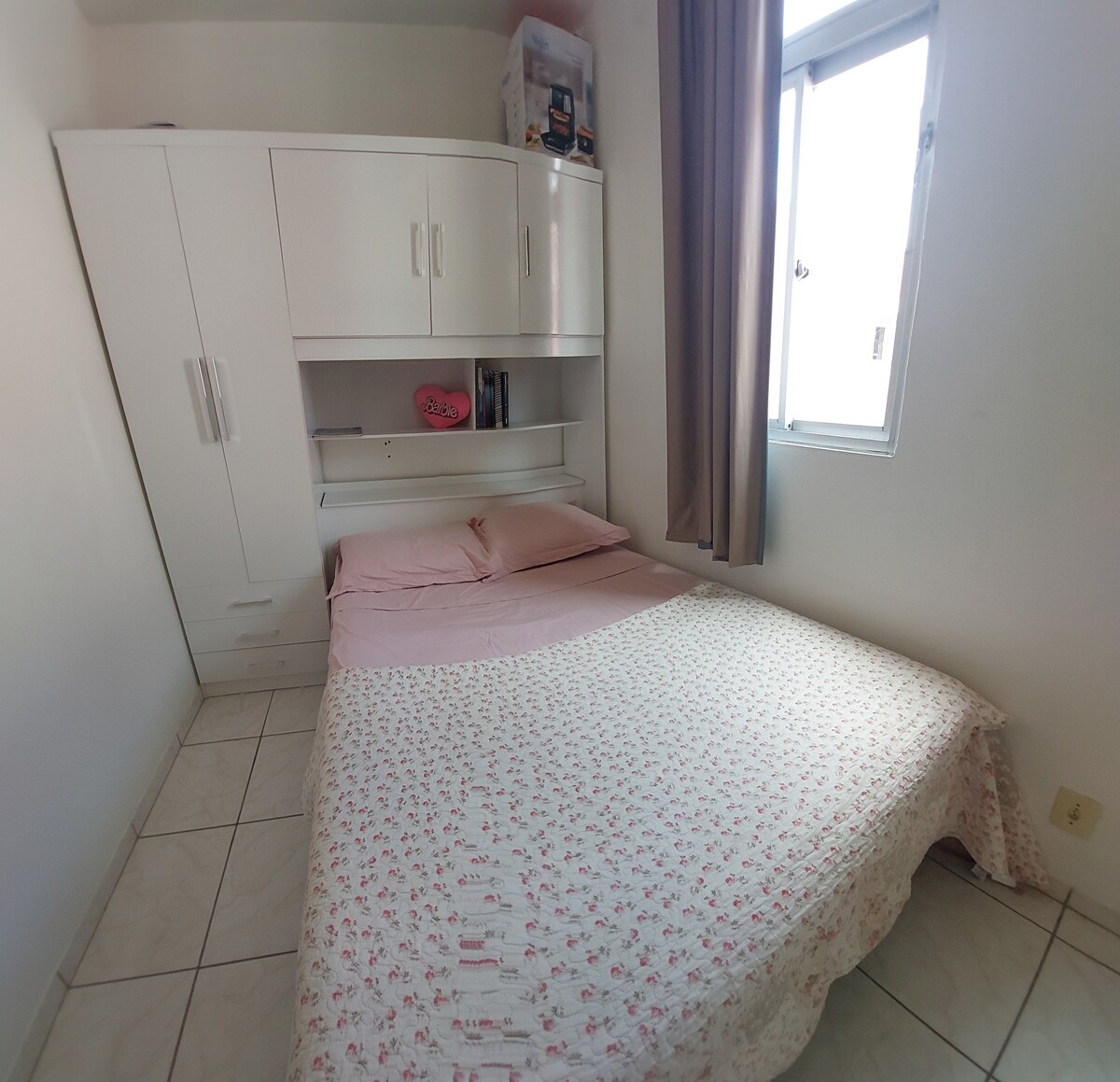 A cozy sleeping area features a spacious bed with a light floral-patterned coverlet and soft pink bedding. A built-in wardrobe provides storage above the bed. Natural light enters through a window adorned with light curtains, enhancing the simplicity of the room.