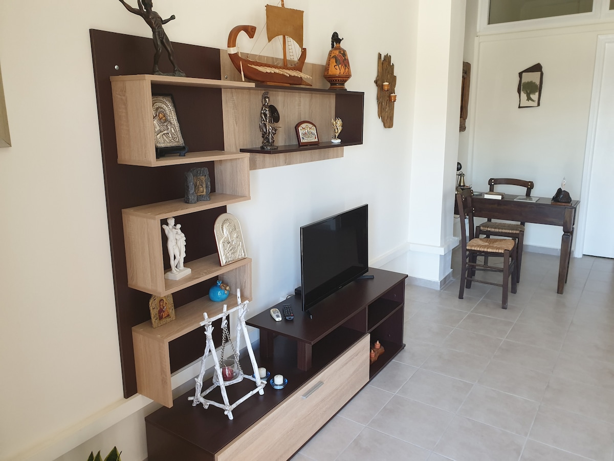 A living area features a modern entertainment unit with decorative items and a television. Light-colored walls complement the open space, which is filled with natural light. A wooden dining table with chairs is visible in the background, enhancing the room's functionality.