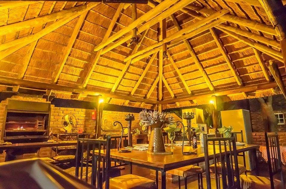 A spacious communal kitchen area is highlighted by a modern thatched roof, featuring wooden beams and a rustic stone wall. A large dining table is surrounded by black chairs, with a centerpiece of decorative plants. An outdoor braai area is visible in the background, providing additional cooking options.