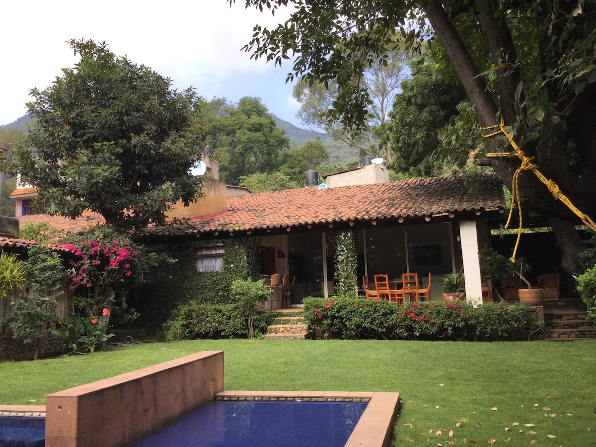 A single-story house is set against a backdrop of lush greenery and mountains. The property features a private garden with vibrant flowers and a pool. Outdoor dining is available with wooden chairs and a table visible in front of the house, enhancing the inviting outdoor space.