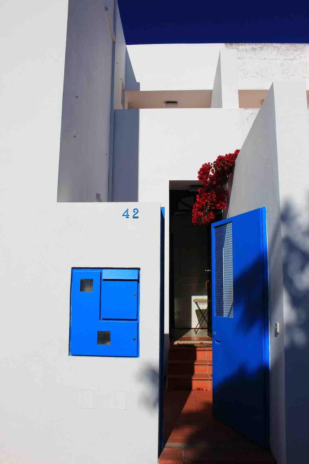 A white exterior leads to an inviting entrance, marked by a bright blue door. A decorative mailbox is visible on the wall beside the door. Vibrant red flowers enhance the entrance, contributing to the overall charm of the entryway.
