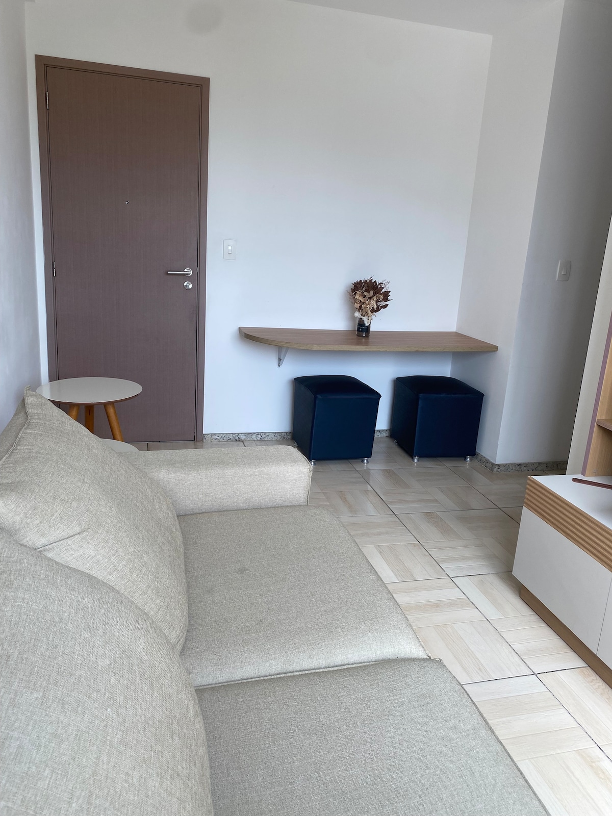 A light, modern living area is shown, featuring a beige sofa and a small round side table. A wooden table with two blue stools is positioned against the wall, complemented by a decorative vase holding dried flowers. Light-colored flooring enhances the space.