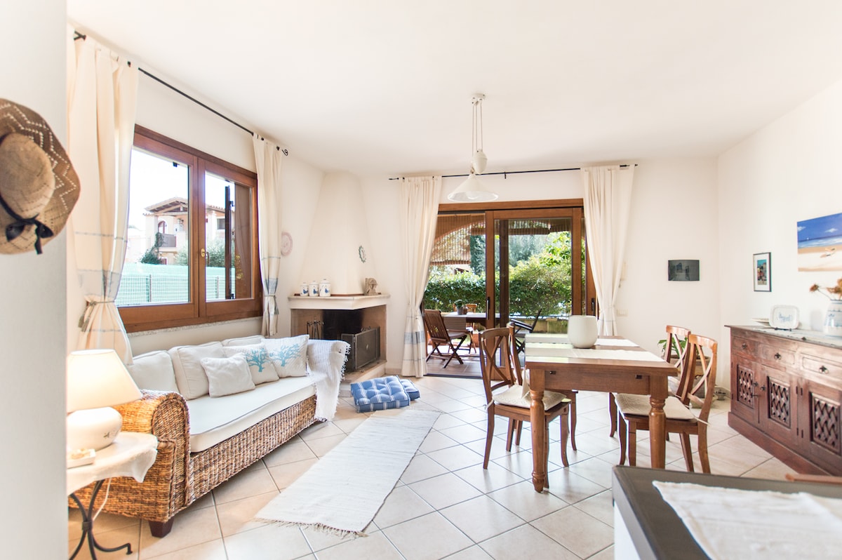 An open living space is showcased with natural light streaming through large windows. A comfortable sofa, wooden dining table, and a decorative sideboard are visible. The area leads to a garden view, framed by soft curtains and a neutral color palette.
